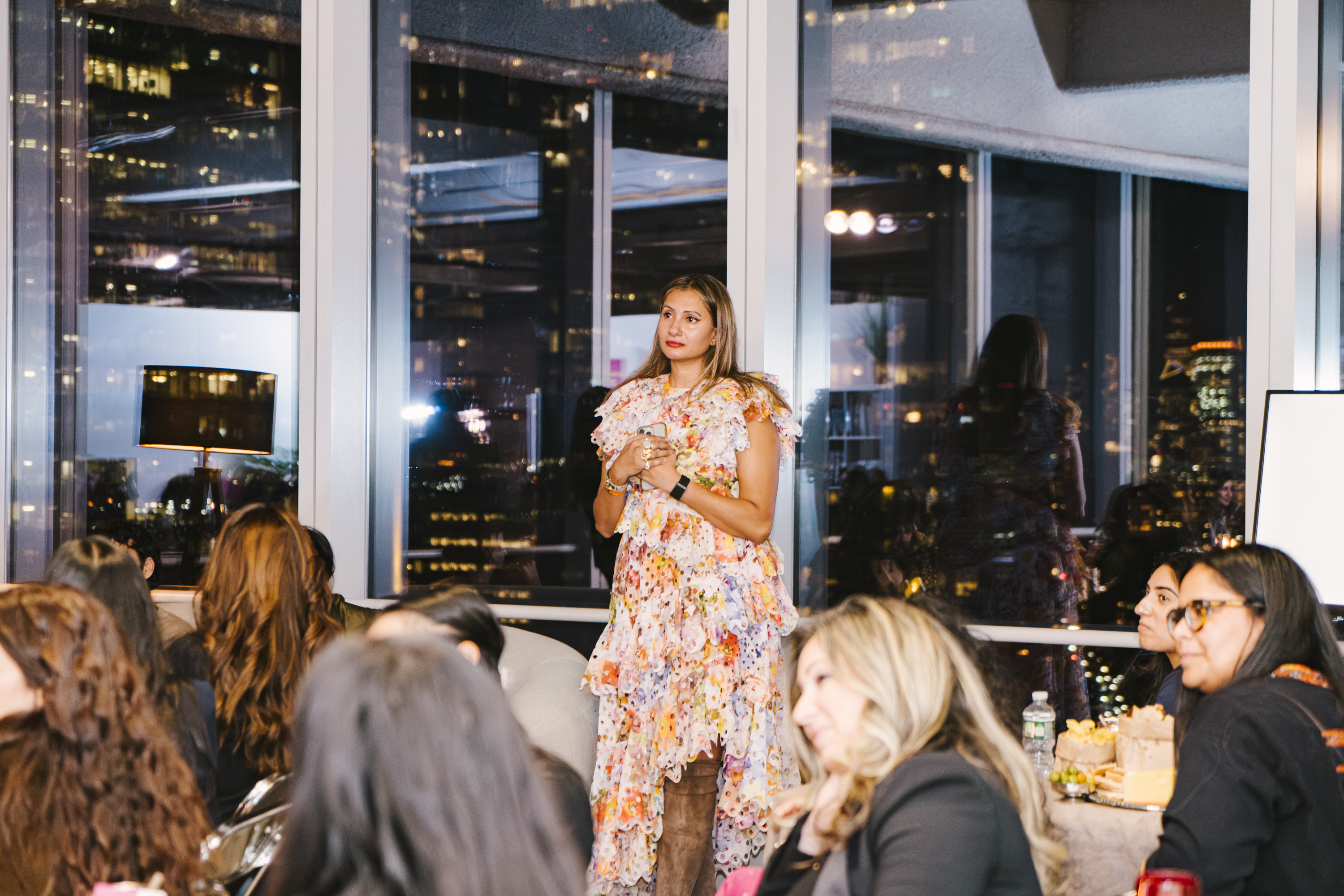 Bhargavi Varma mid-room at the Brown Girl Angels SickKids fundraiser in New York, NYC skyline behind her — the founder addressing the room.