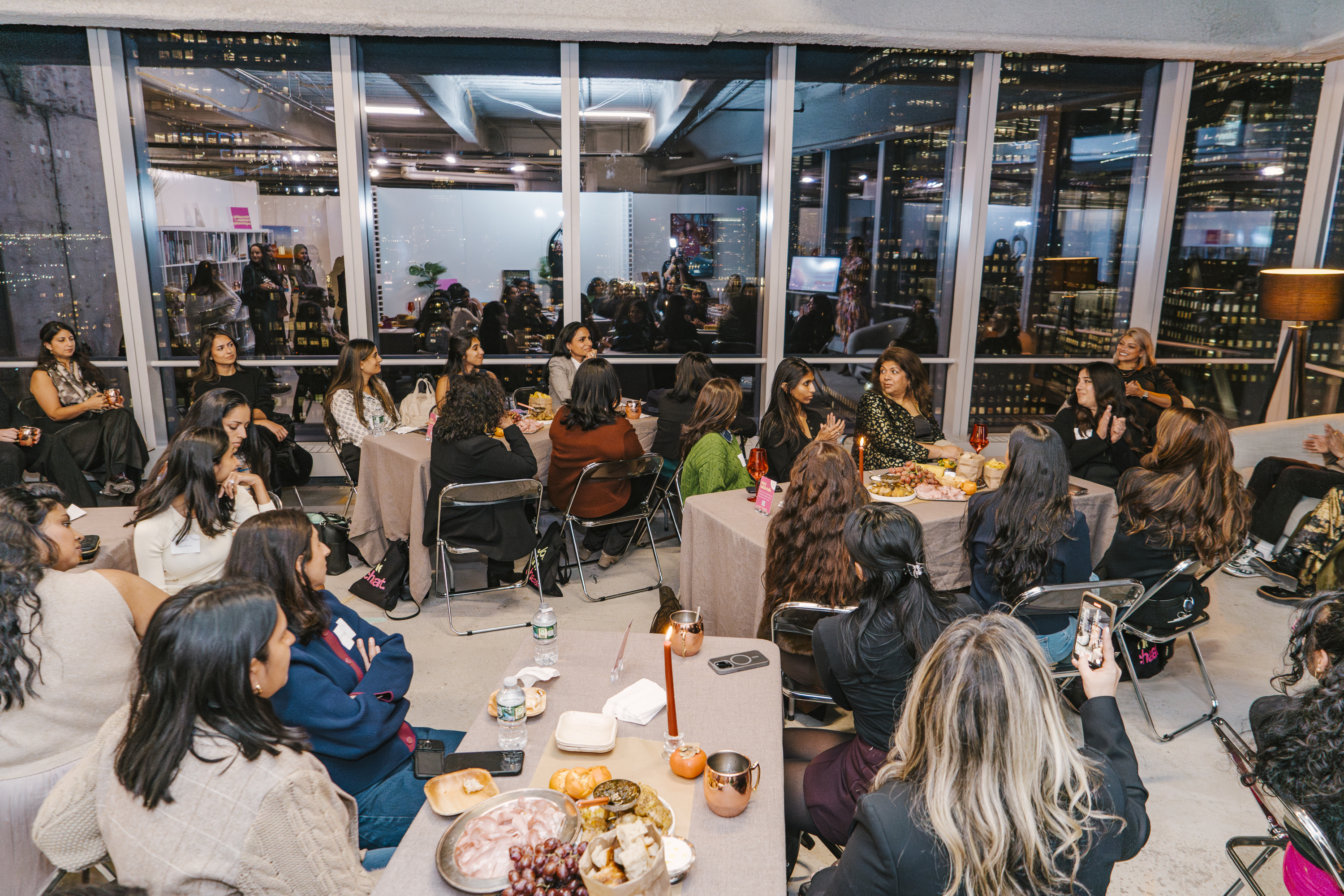 A wide shot of a Brown Girl Angels community dinner in New York &mdash; the long table, the skyline, the room a Guest Seat opens you into.